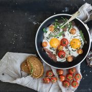 Pan of fried eggs, bacon and cherry-tomatoes with bread on dark table surface, top view