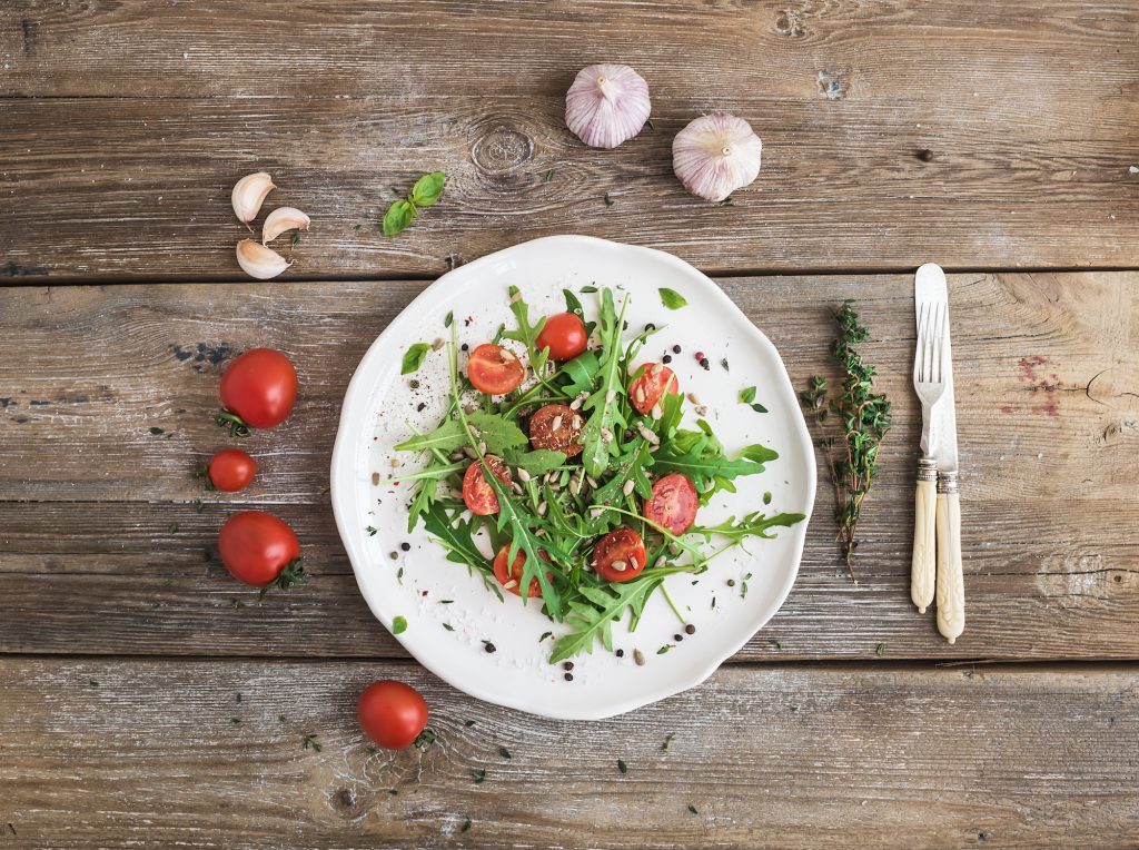 Salad with arugula, cherry tomatoes, sunflower seeds and herbs on white ceramic plate over rustic wood background, top view, copy space