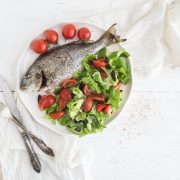 Cooked sea bream fish with fresh vegetable salad on ceramic plate over white rustic wooden backdrop, top view, copy space